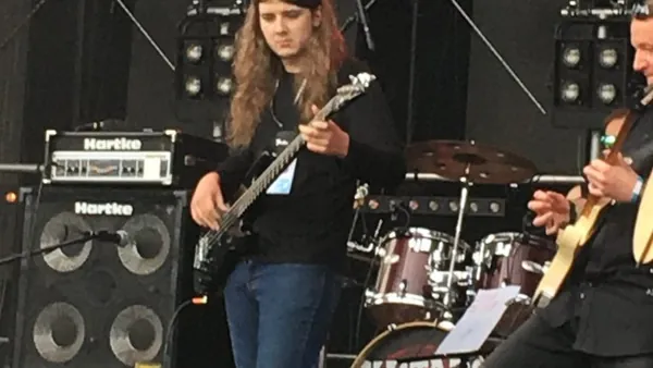Mateusz Pawelec z gitarą basową na plenerowej scenie, za nim wzmacniacze i perkusja