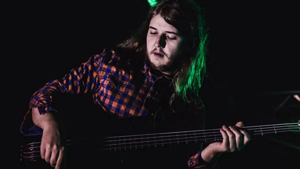 Mateusz Pawelec w koszuli w kratę gra na gitarze basowej w półmroku