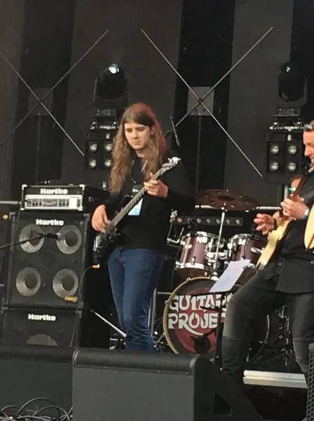 Basista z długimi włosami gra na scenie, za nim wzmacniacze i perkusja