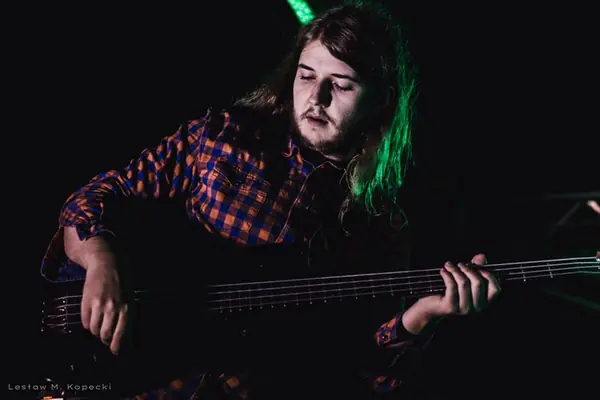Mateusz Pawelec w koszuli w kratę gra na gitarze basowej w półmroku